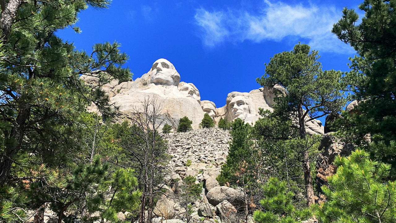 A view of the Mount Rushmore carvings framed by green pine trees from a hiking trail perspective.